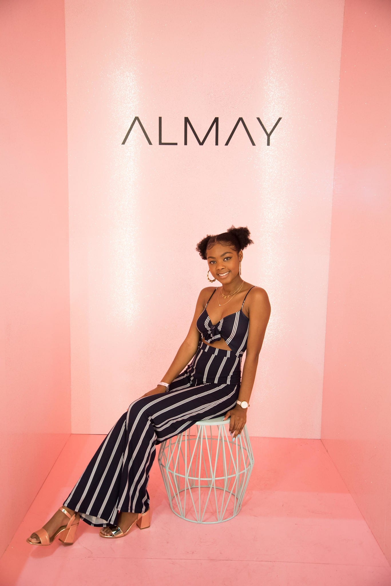 A guest in a striped jumpsuit seated on a wire stool against the pink ALMAY backdrop at the beauty activation