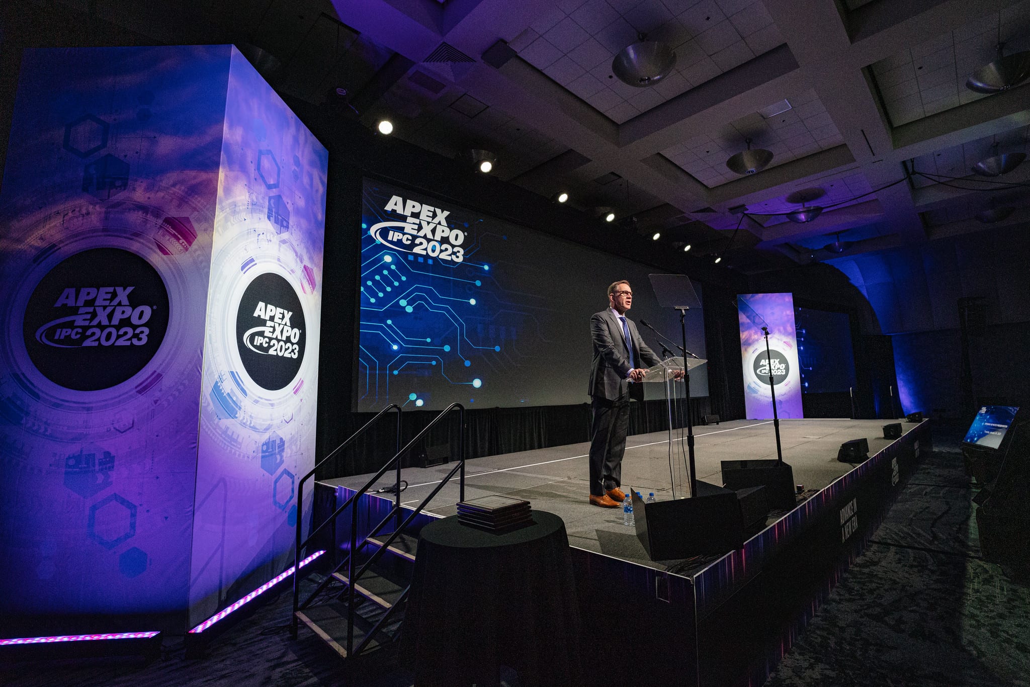 A speaker at the podium on a purple-lit main stage under the IPC APEX EXPO 2023 branding