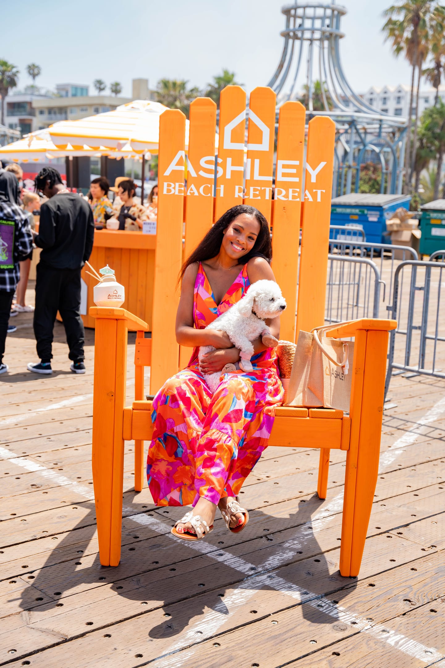 A guest in a bright floral dress holding a small white dog in a giant orange Ashley-branded chair at a beachside brand activation on the Santa Monica pier
