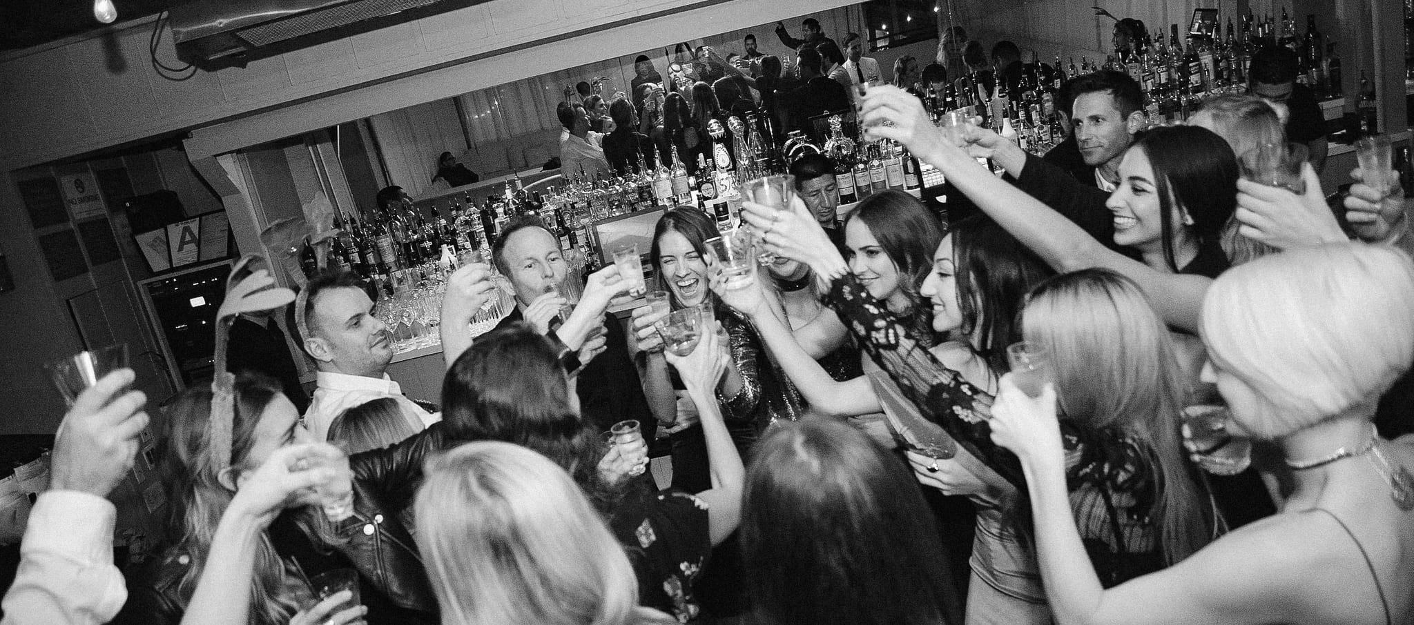 Guests raising glasses in a toast at the bar of the BELLA+CANVAS holiday party, in black and white