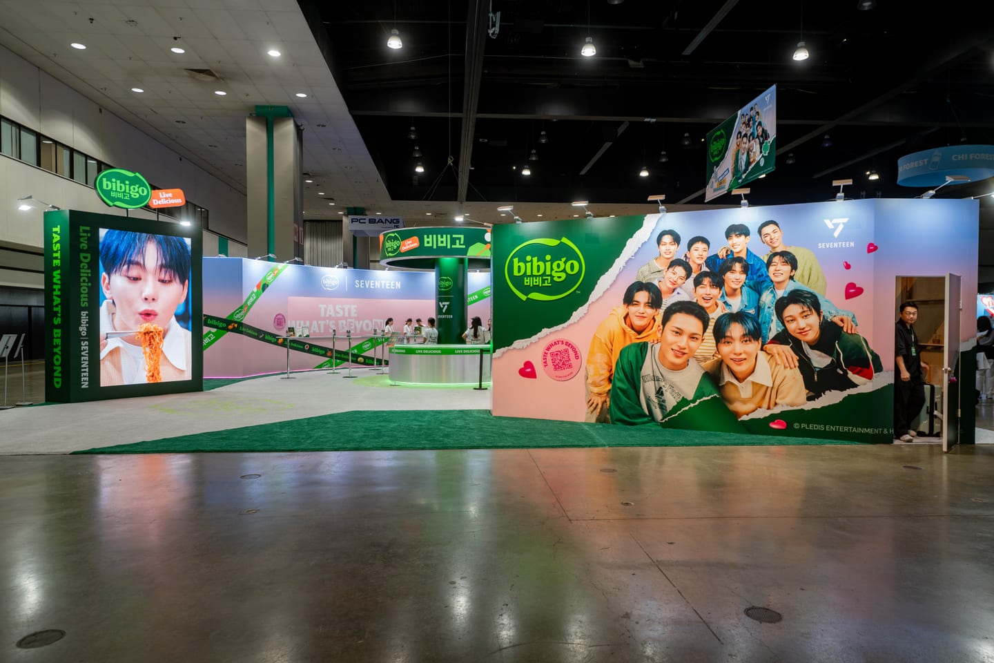 The green bibigo booth at KCON LA 2025 branded with the K-pop group SEVENTEEN, showing a torn-paper mural of the members and a Taste What’s Beyond LED wall
