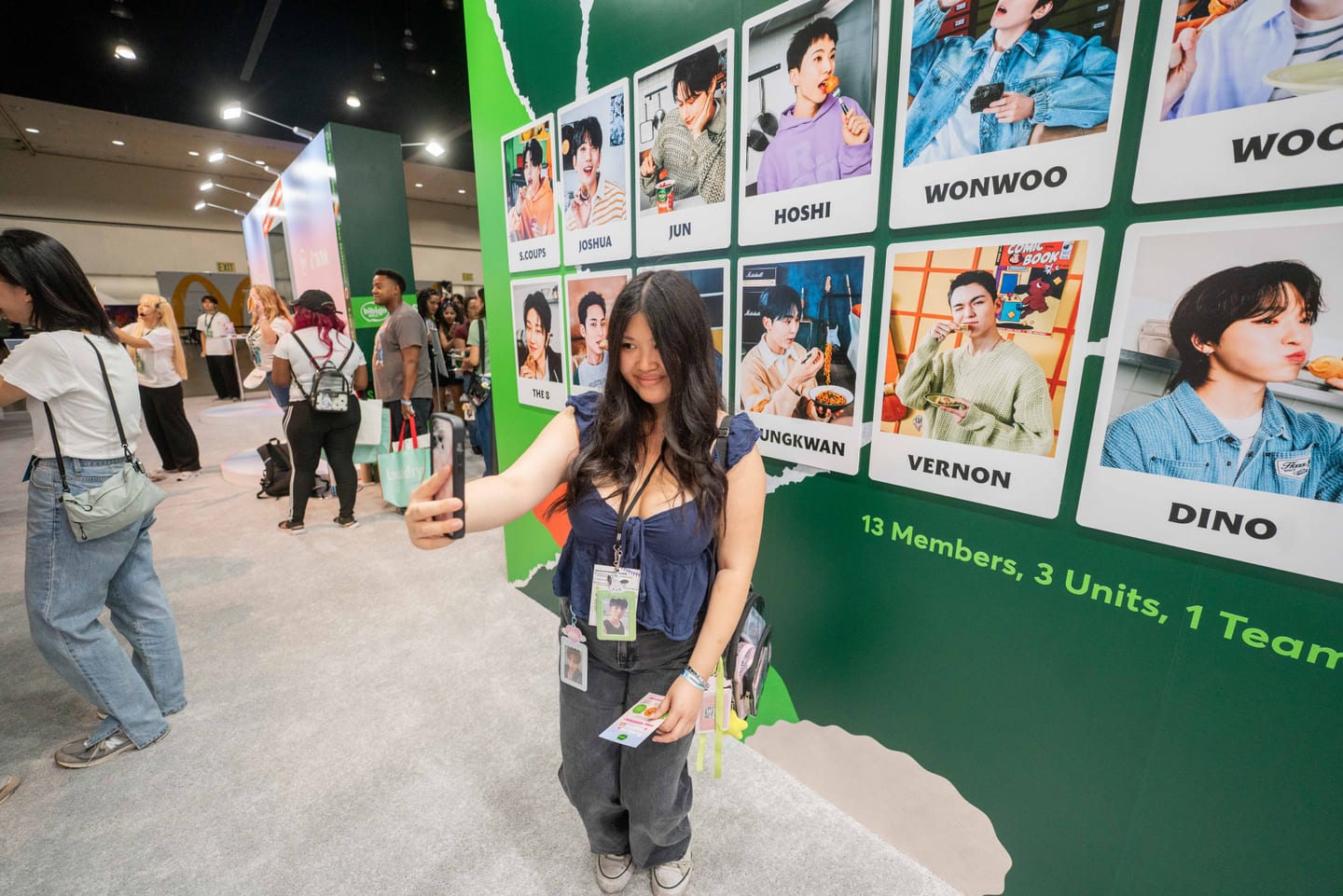 A fan taking a selfie in front of the green bibigo photo wall of all thirteen SEVENTEEN members at KCON LA 2025