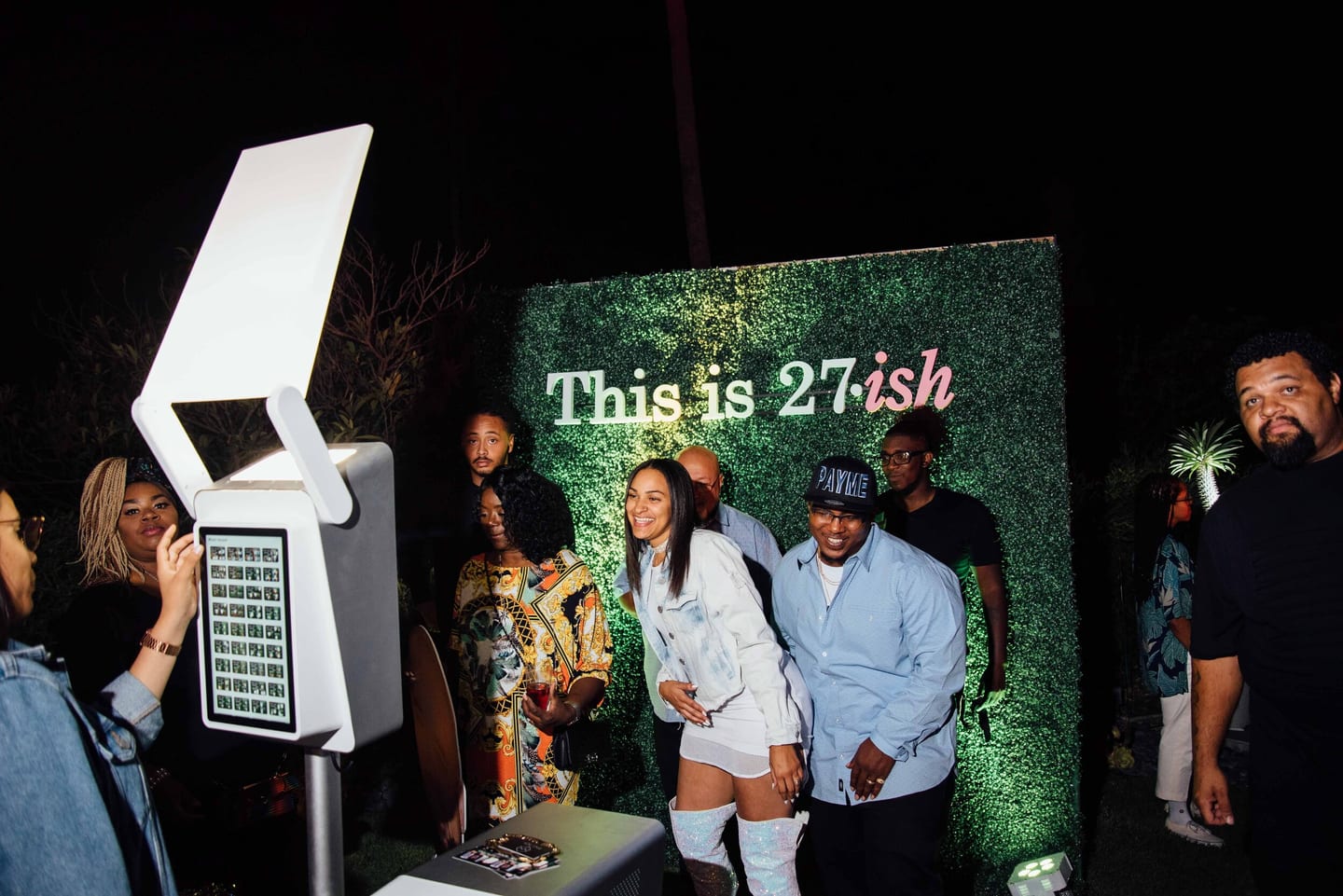 Guests posing at a photo booth with a This is 27-ish backdrop at a birthday party