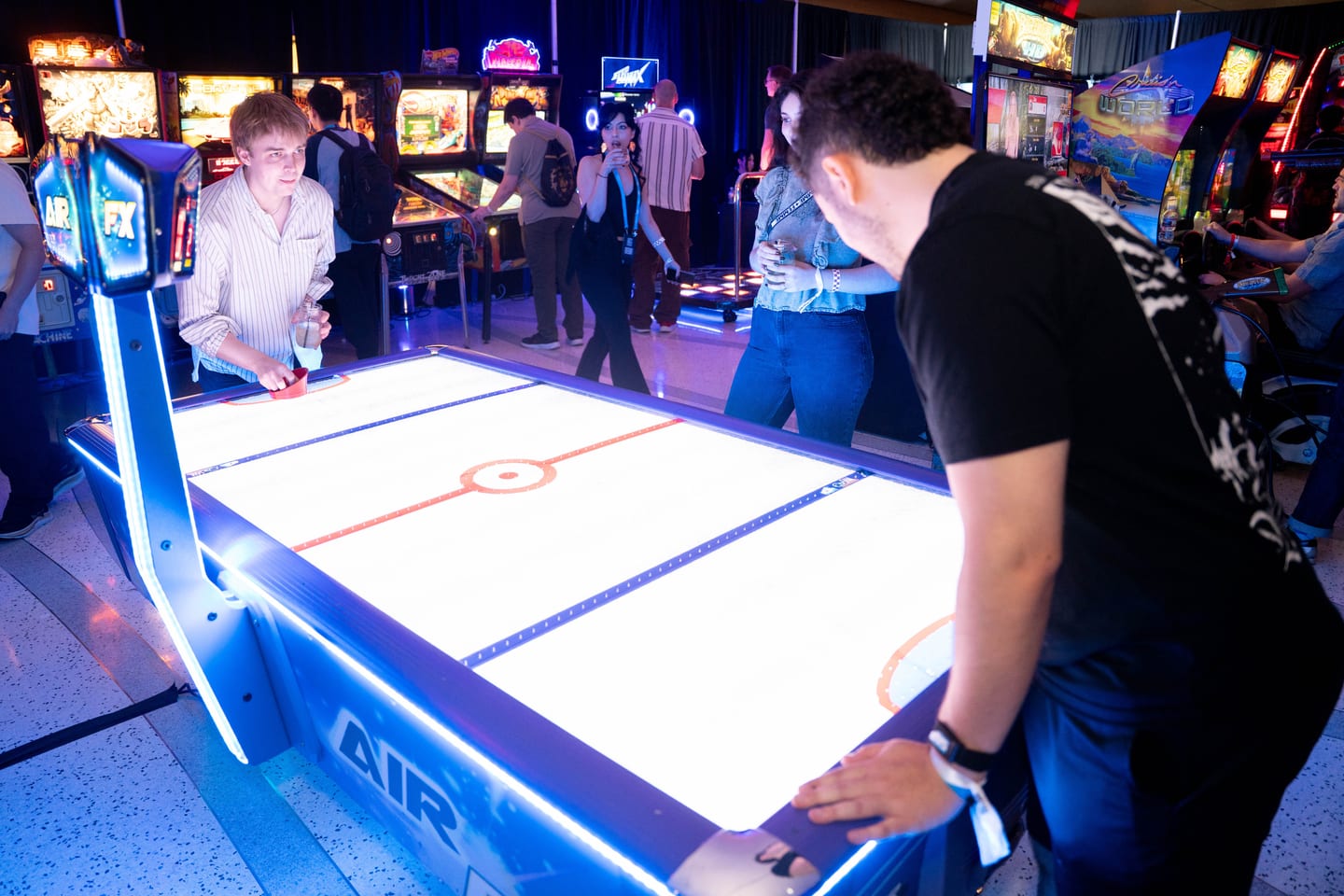 Two guests playing a glowing LED air hockey table in the arcade, with pinball machines behind them