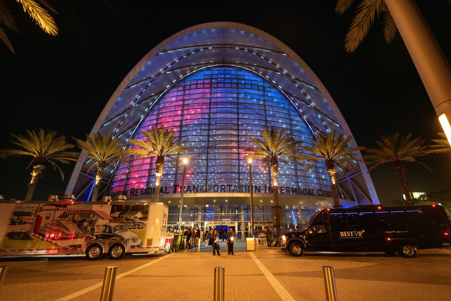 The arched facade of the Anaheim Regional Transportation Intermodal Center lit pink and blue at night, with food trucks parked out front