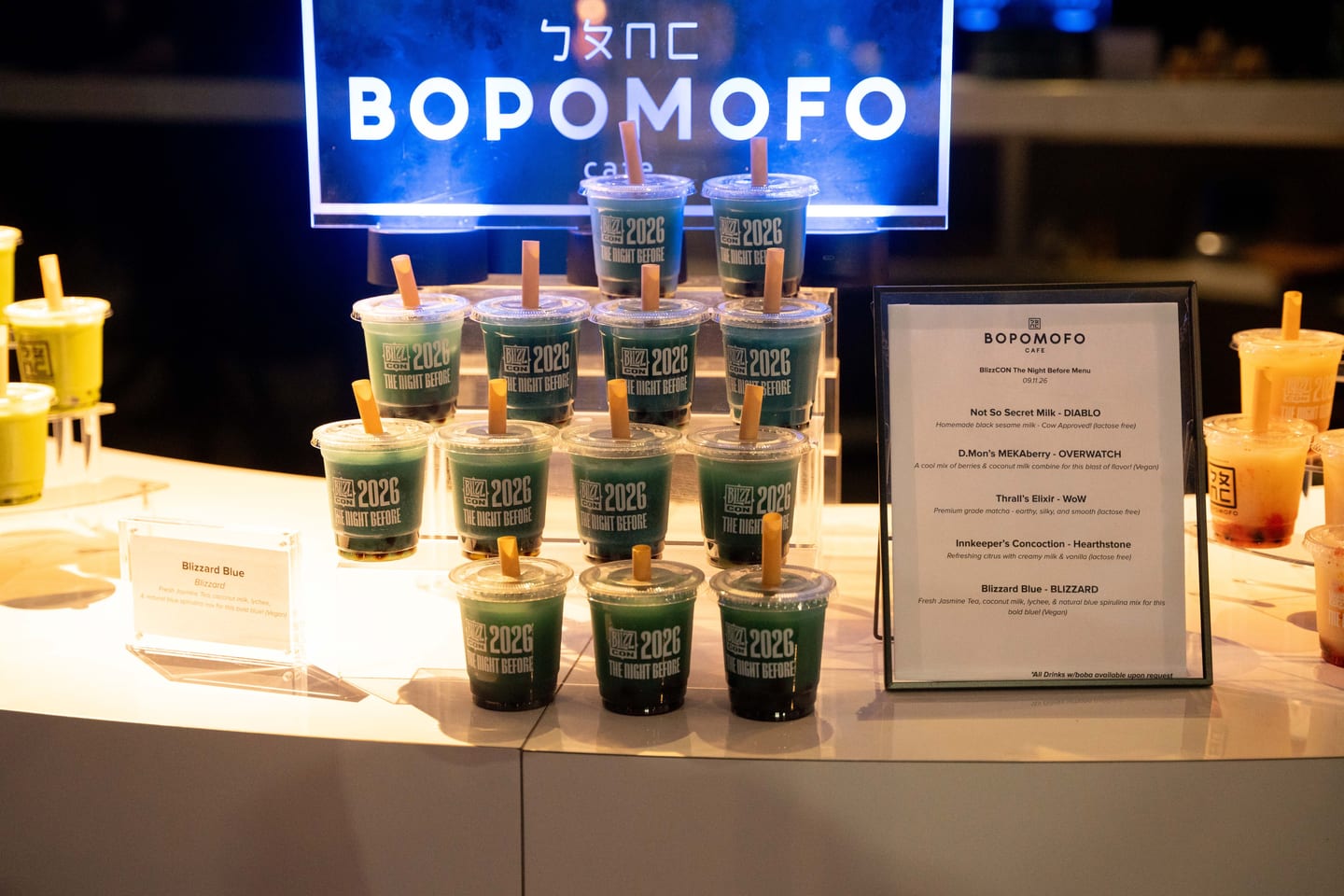 Rows of blue boba drinks in BlizzCon 2026 The Night Before cups beside a Bopomofo Cafe menu of drinks named after Blizzard games