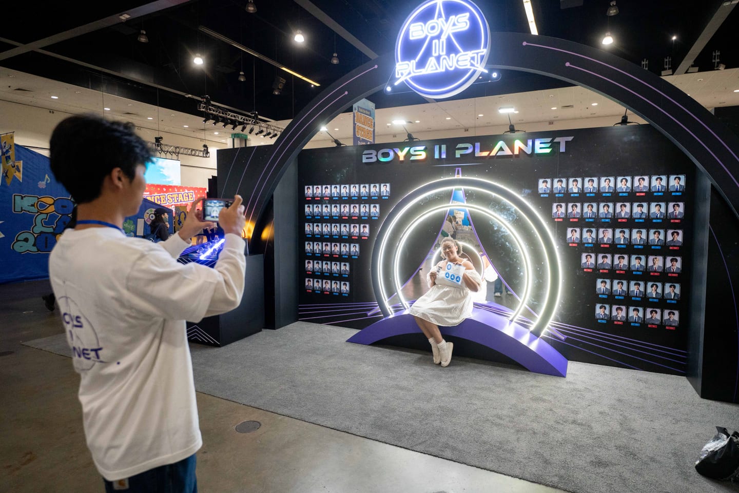 A fan being photographed on the lit Boys II Planet stage at KCON LA 2025 in front of a wall of contestant portraits