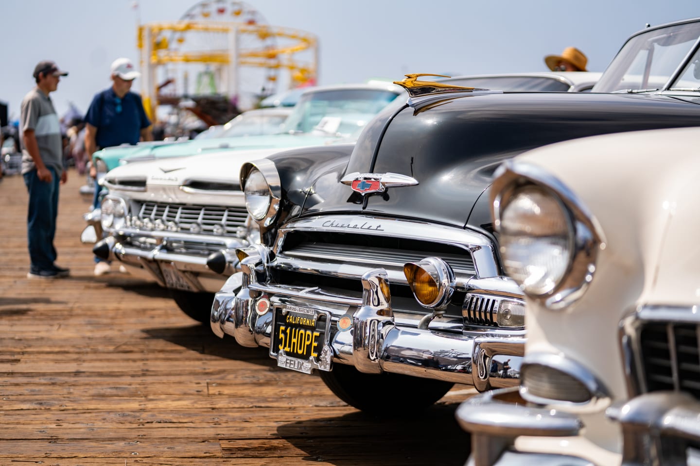 A row of polished 1950s Chevrolets with chrome grilles at a classic car show on the Santa Monica Pier, the Pacific Park ferris wheel behind