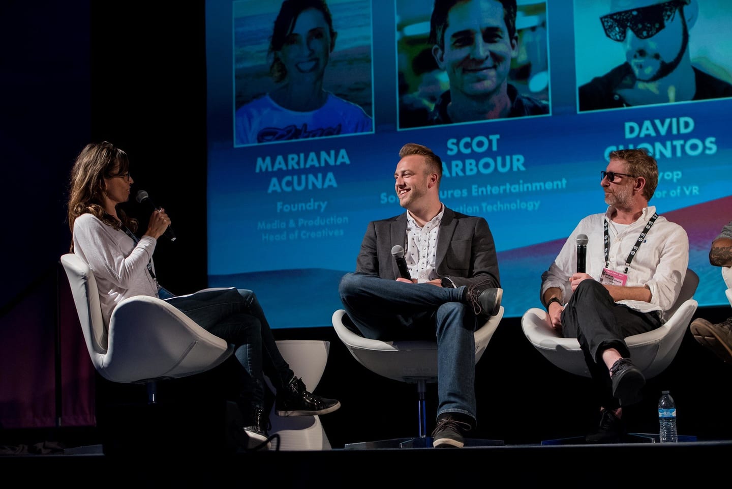 Three-person speaker panel on stage with speaker headshots projected behind at a conference