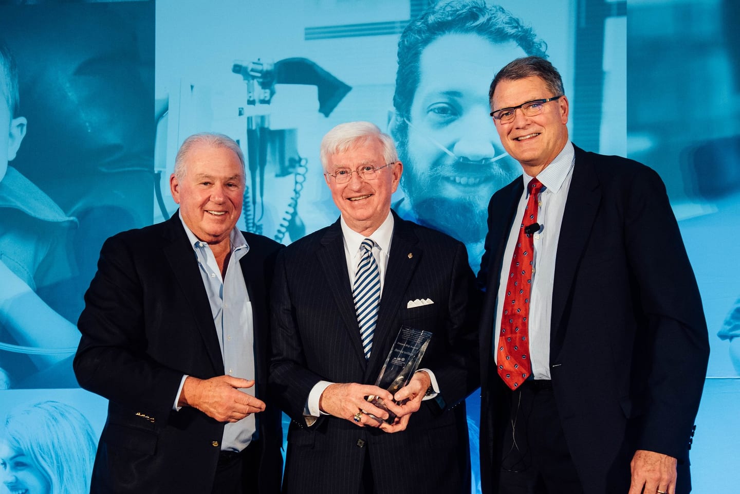 Three executives on stage as an honoree holds a glass award trophy at a corporate awards event