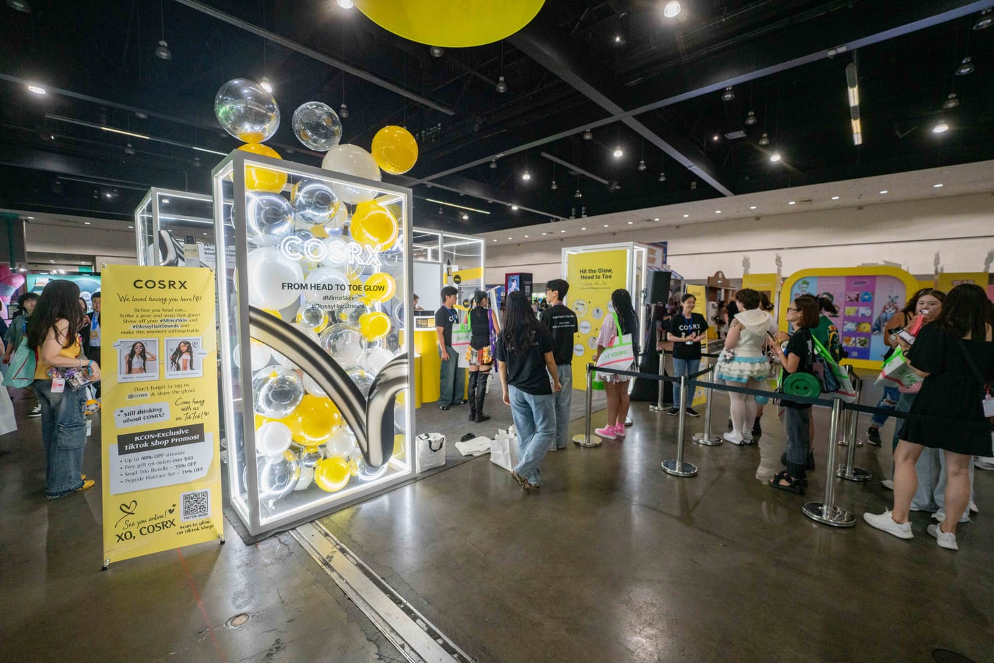 The yellow and white COSRX booth at KCON LA 2025 with a From Head To Toe Glow bubble display and a queue behind stanchions