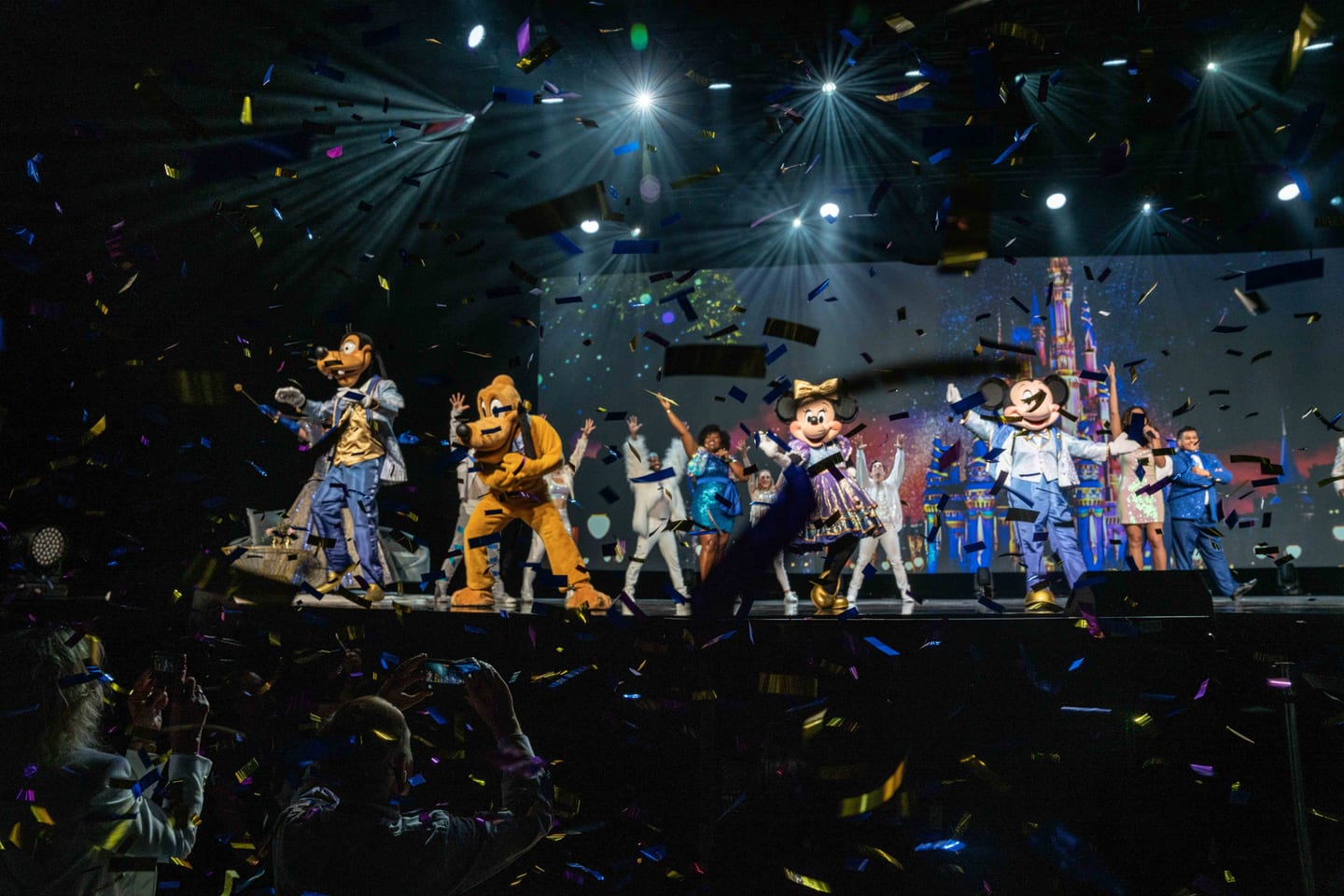 Mickey, Minnie, Pluto and Goofy performing on stage under confetti with a castle backdrop at the IPW travel trade show showcase in Orlando
