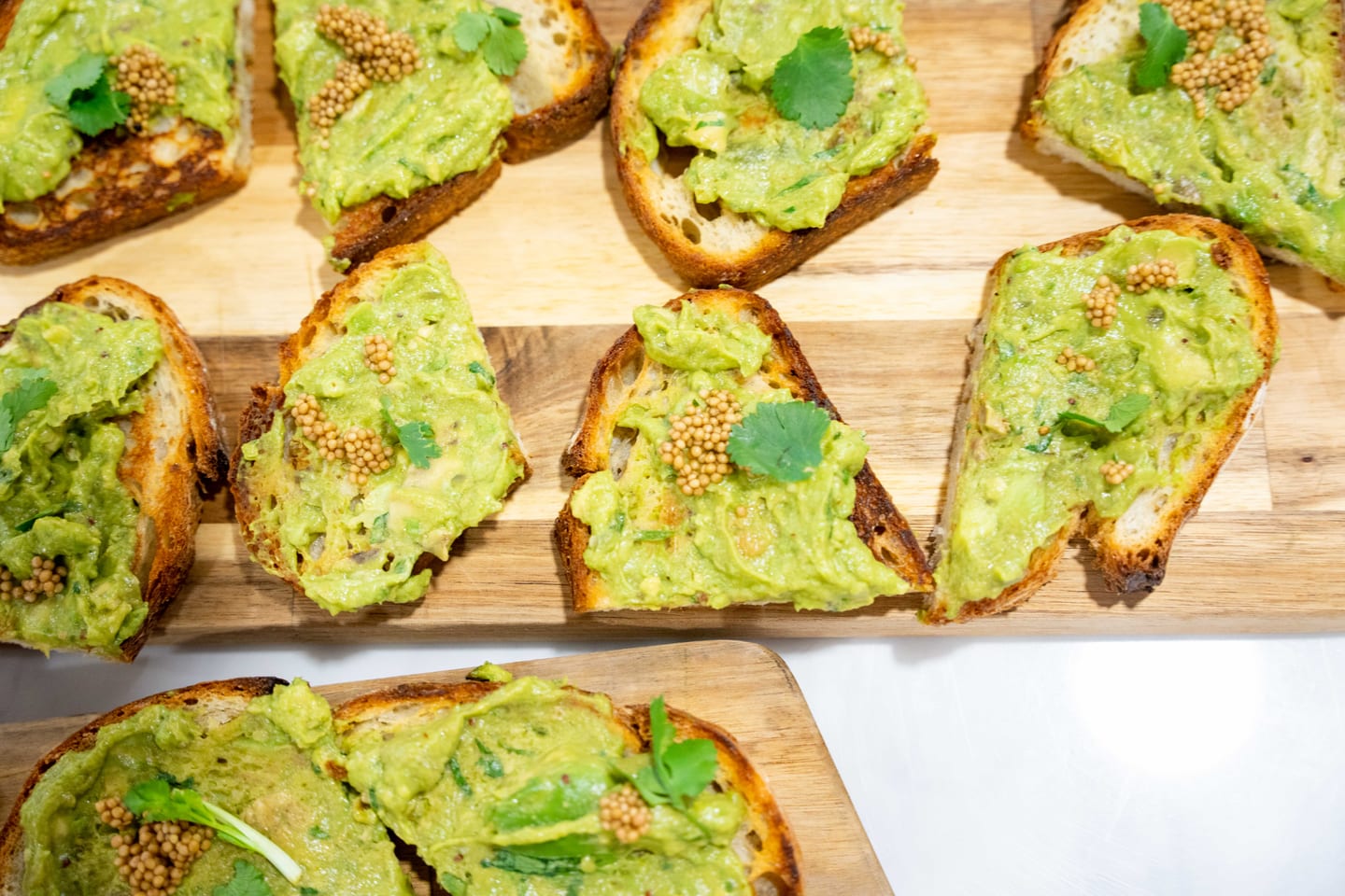 Overhead view of avocado toast points on a wooden board, garnished with pickled mustard seeds and cilantro