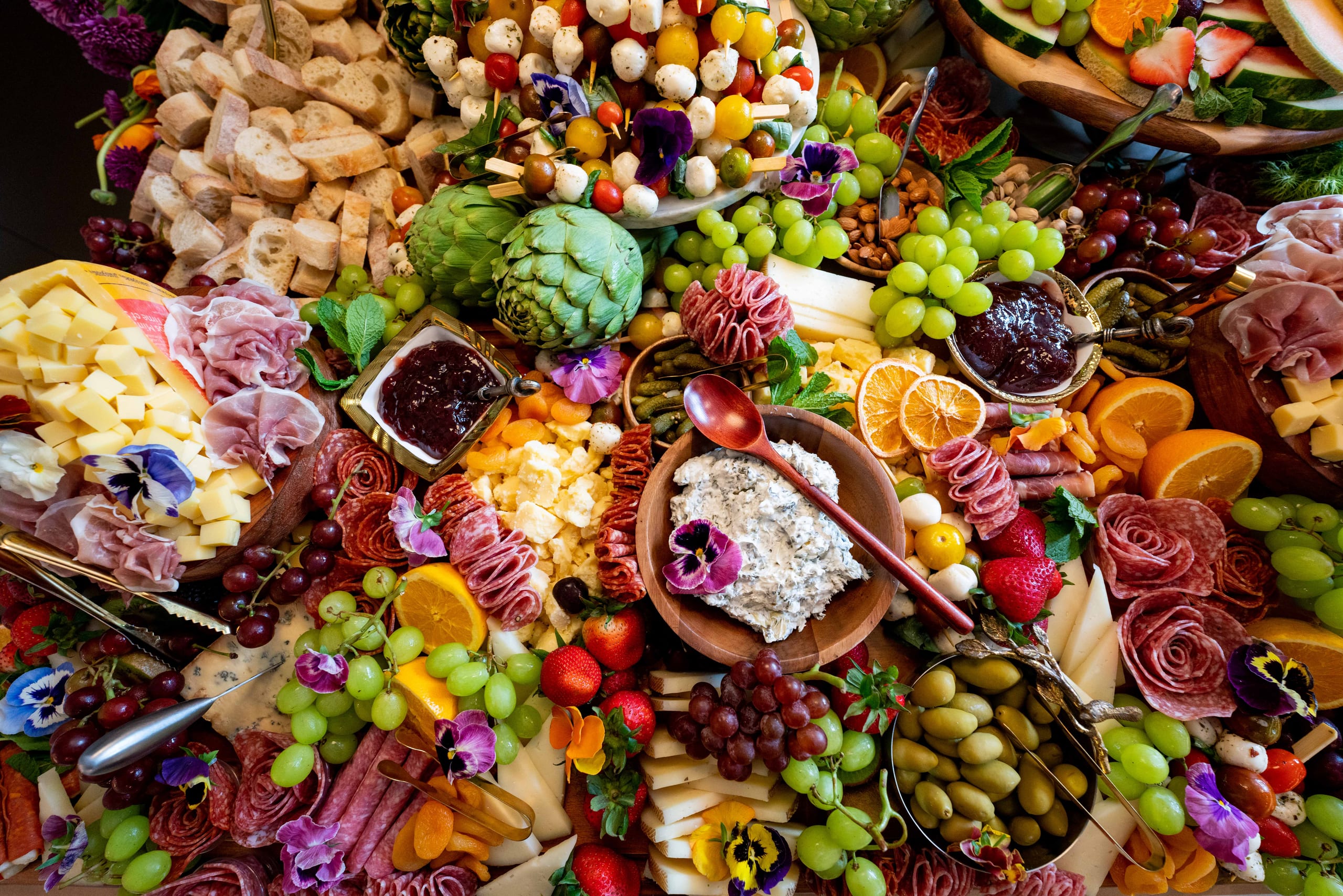 Overhead view of an abundant grazing table with charcuterie, cheeses, grapes, olives, dips, sliced fruit, and edible flowers