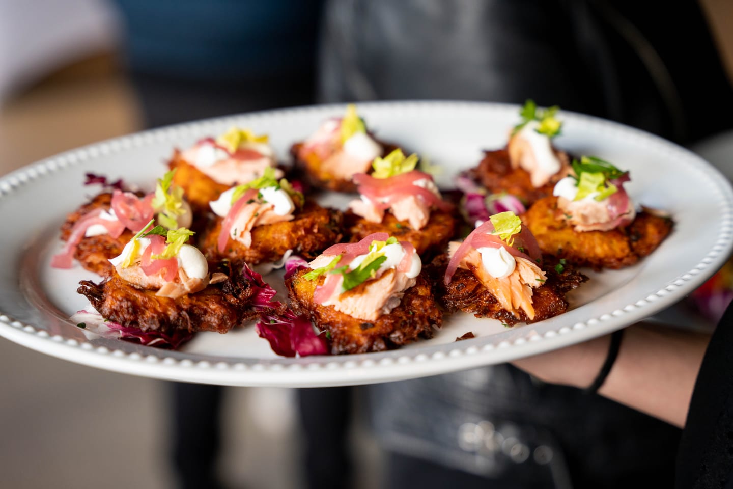 A tray of passed hors d’oeuvres — crisp potato cakes topped with smoked salmon, crème fraîche, and pickled onion — carried by a server