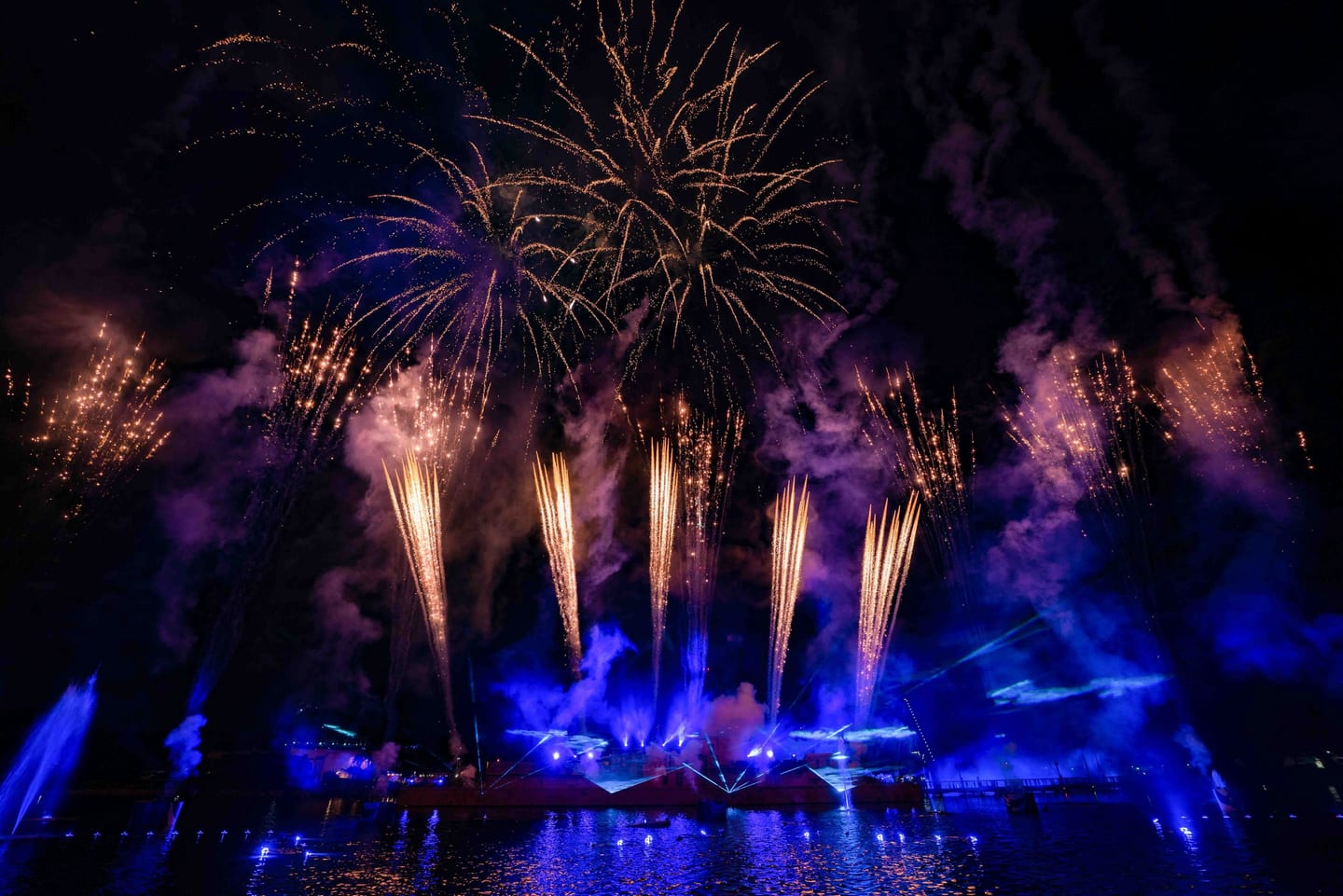 Fireworks and a blue light show reflected over water at a nighttime event finale