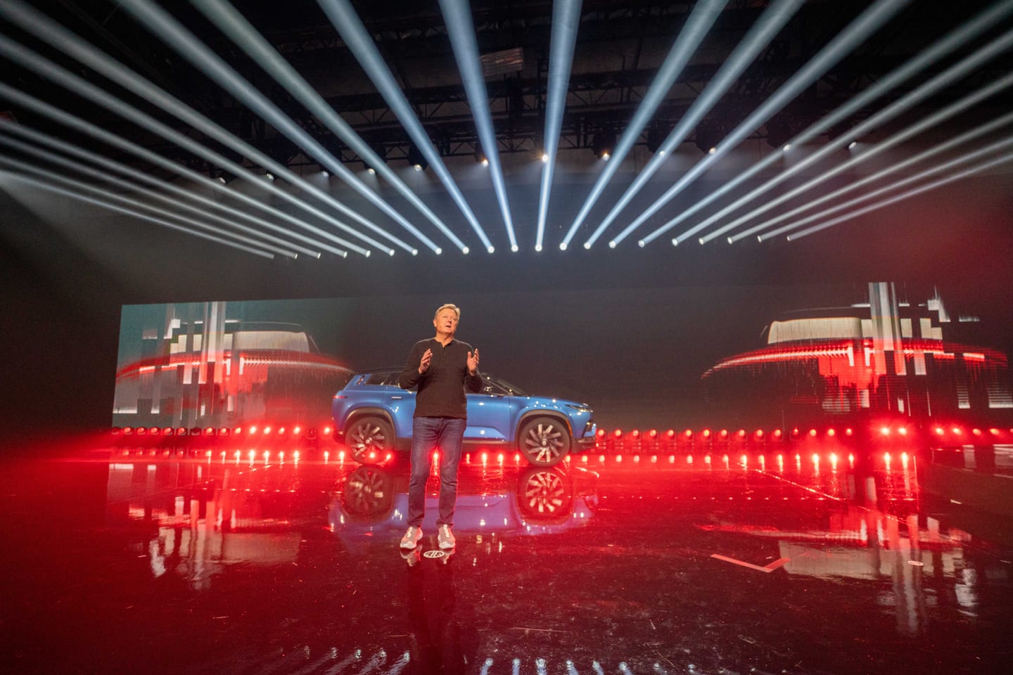 A presenter speaking beside the blue Fisker Ocean under rows of white beams and red floor lights