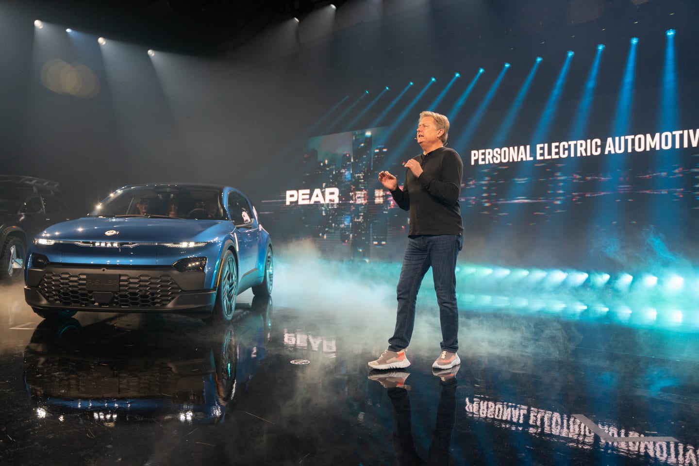 A presenter beside the blue Fisker PEAR, the screen reading Personal Electric Automotive Revolution