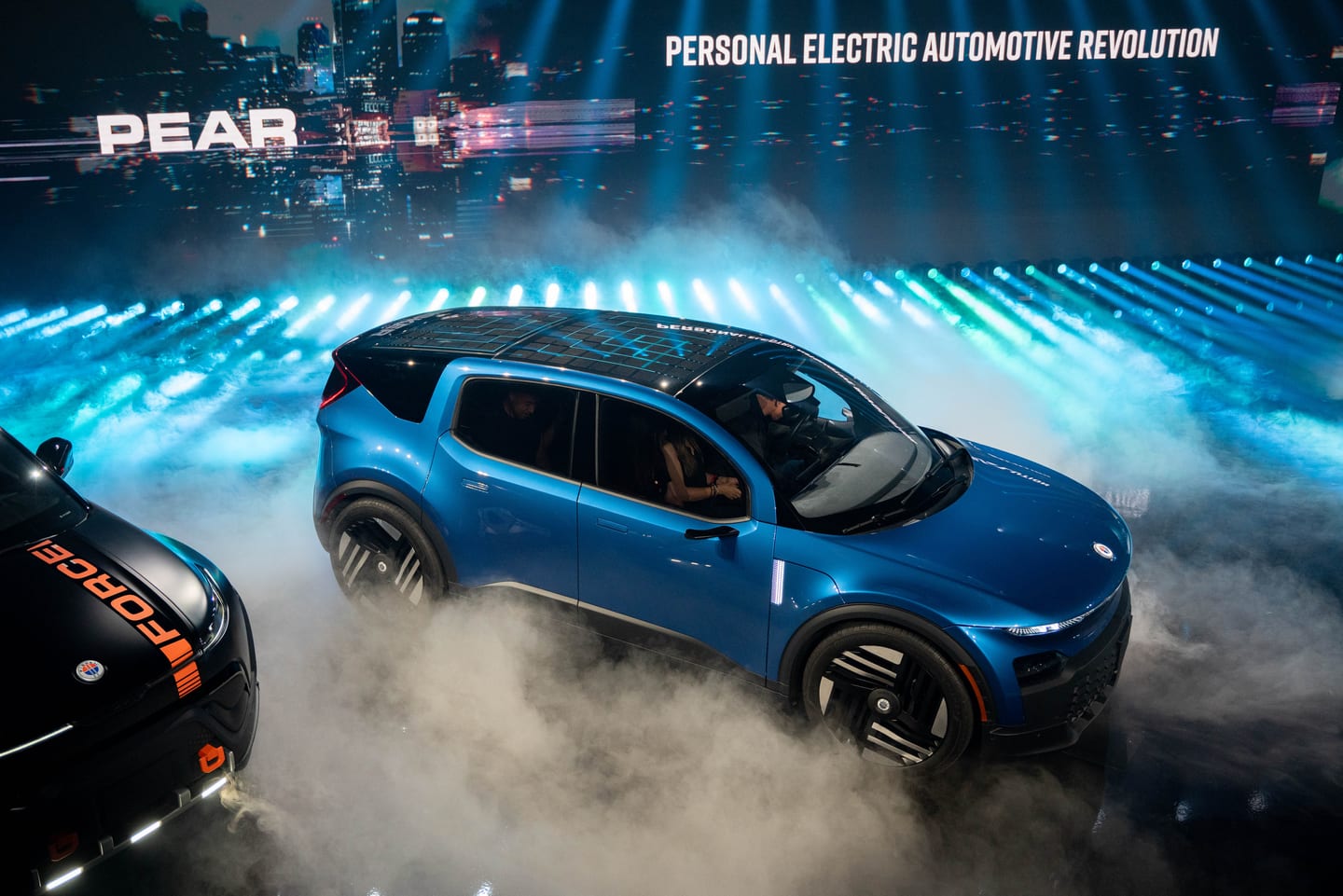 The blue Fisker PEAR moving through stage fog beneath the Personal Electric Automotive Revolution line