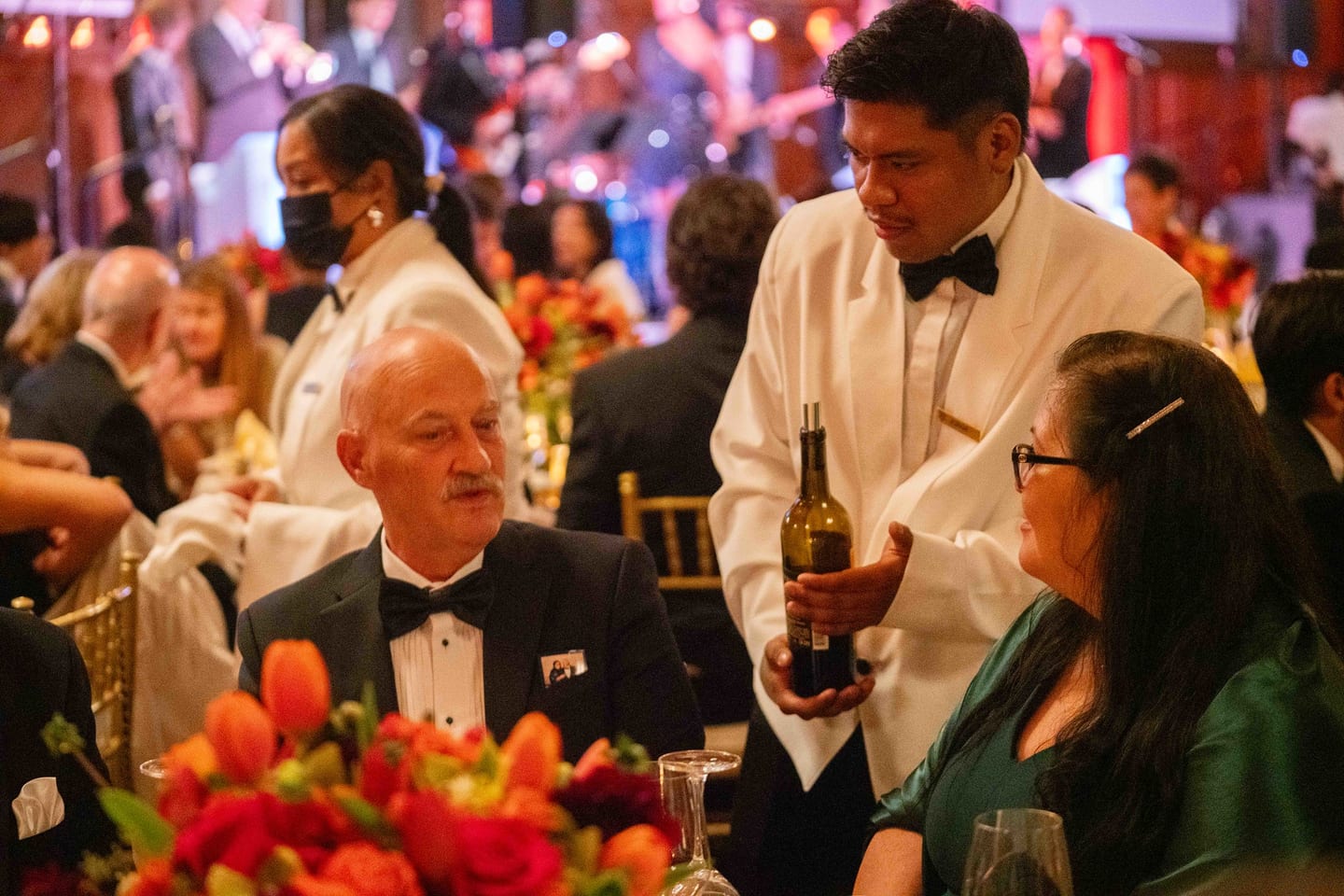 Waiter in a white jacket pouring wine tableside at a gala with a live band on stage behind