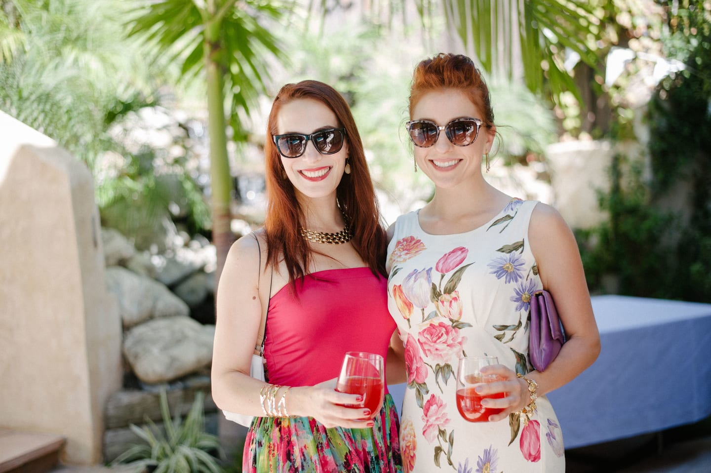 Two guests in sunglasses and floral dresses with drinks at a garden party