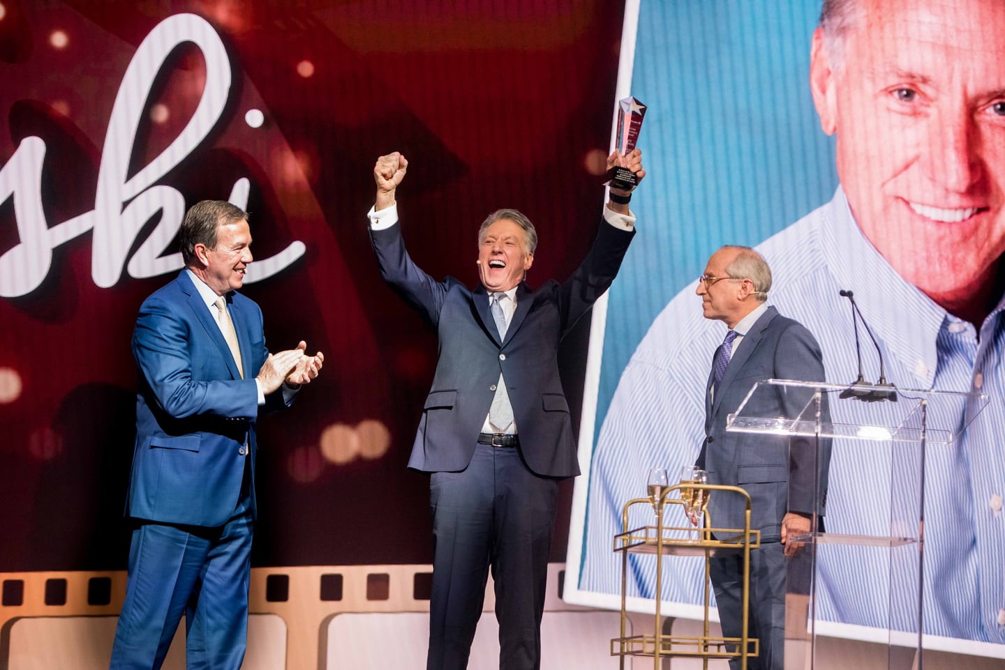 An honoree raising a trophy and fist on stage as a presenter applauds at the Hunter Douglas award gala at the Dolby Ballroom in Hollywood