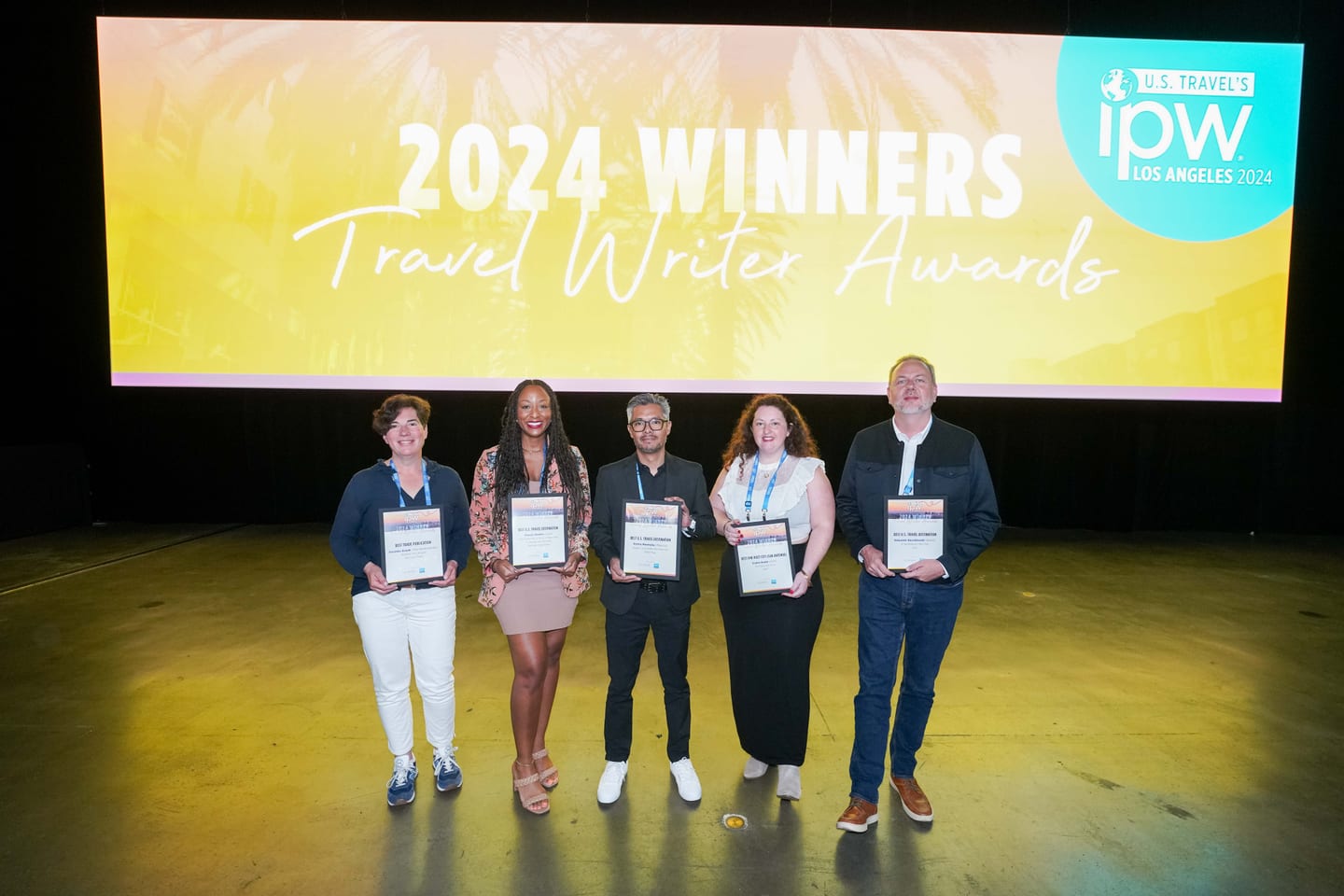 Award winners standing on stage holding certificates beneath a 2024 Winners Travel Writer Awards screen at IPW