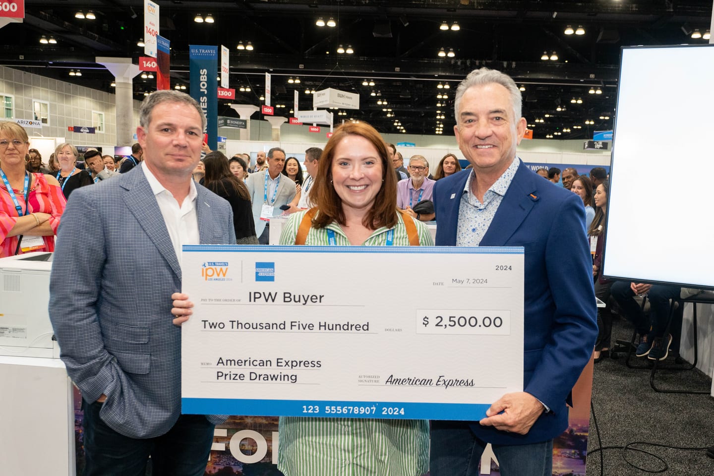 Two people presenting an oversized American Express prize-drawing check for $2,500 to an IPW buyer on the trade-show floor