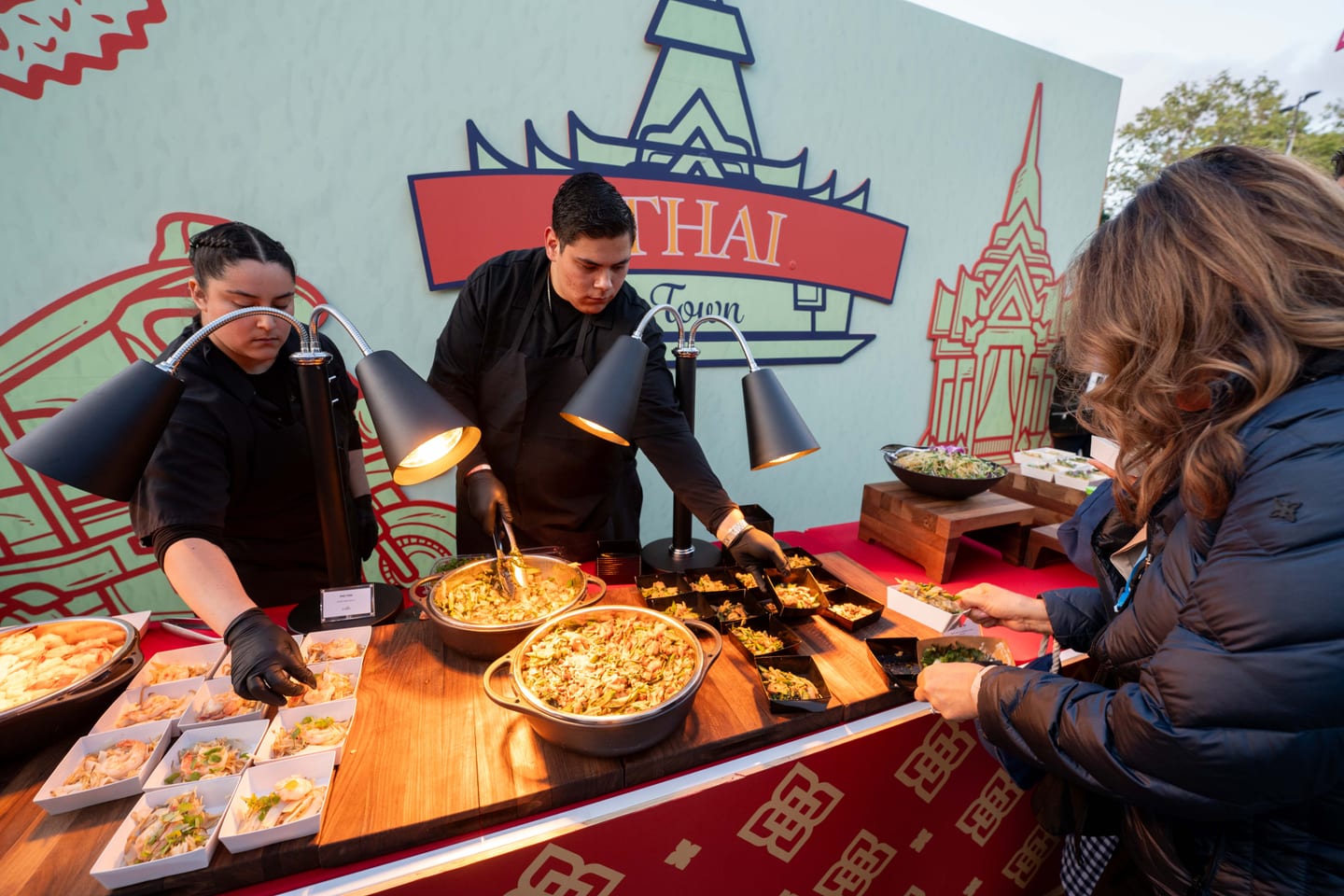 A chef plating food at a Thai culinary station beneath a THAI backdrop as a guest is served at an IPW reception
