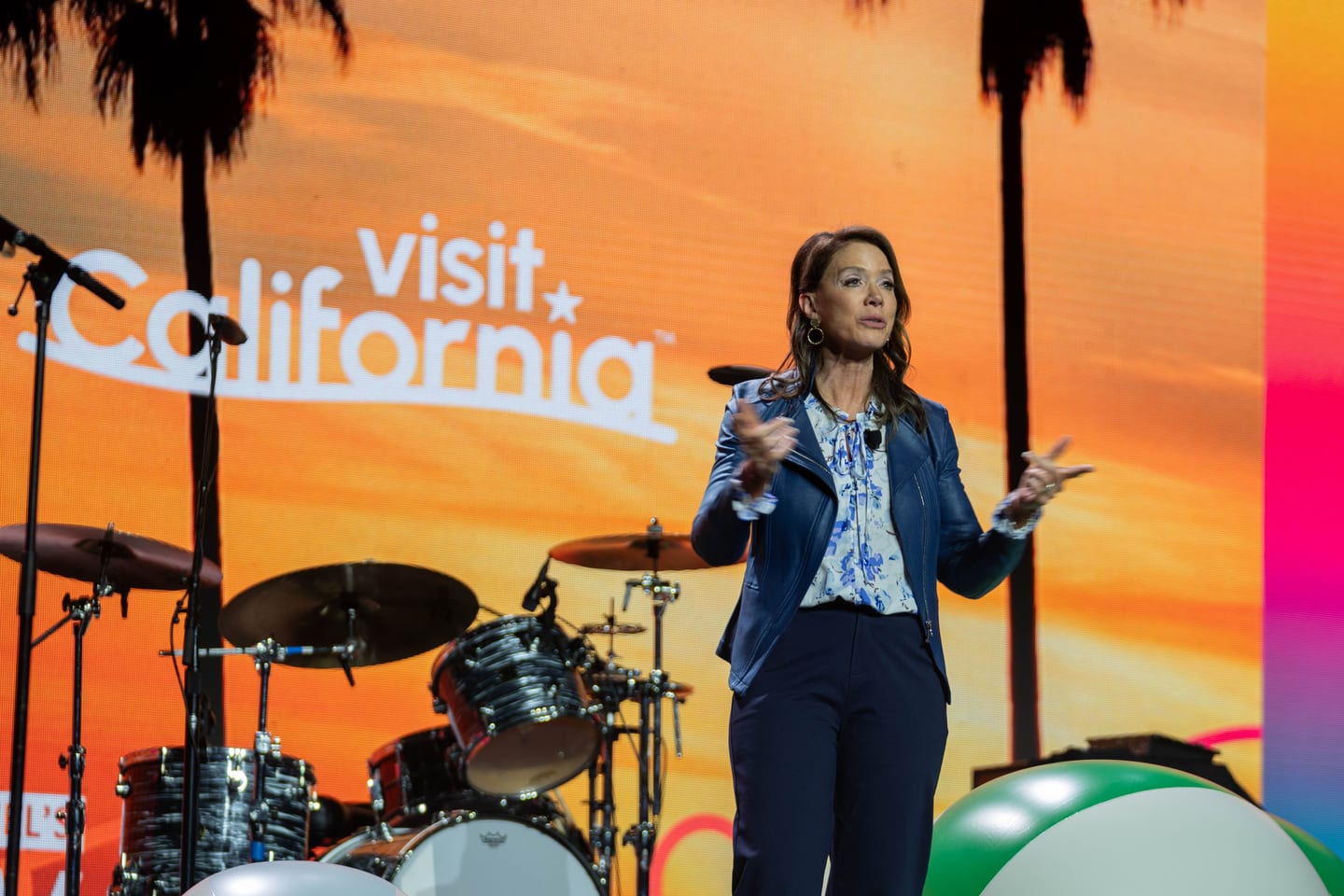 A speaker presenting on the IPW main stage in front of a Visit California sunset screen with a band set up beside the stage