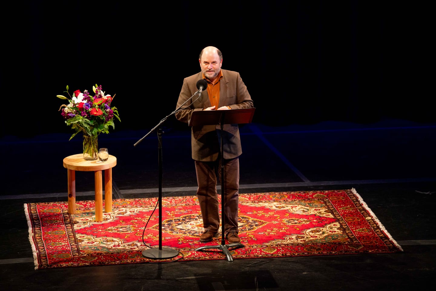 Actor Jason Alexander speaking at a lectern on a small rug-set stage at an event