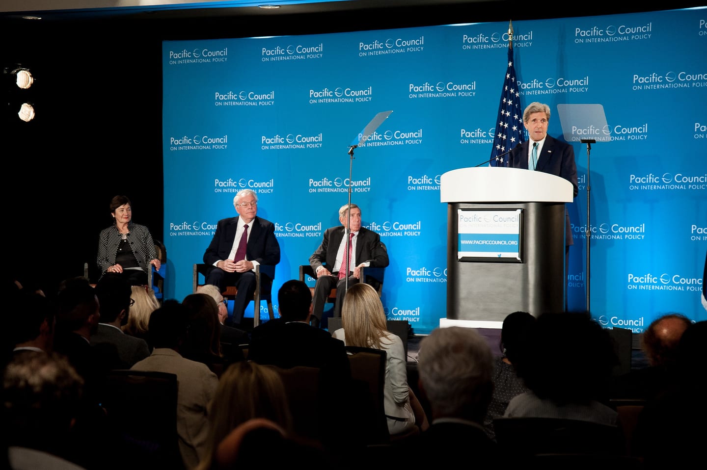 Former Secretary of State John Kerry speaking at a podium with seated panelists and a U.S. flag on stage at the Pacific Council on International Policy