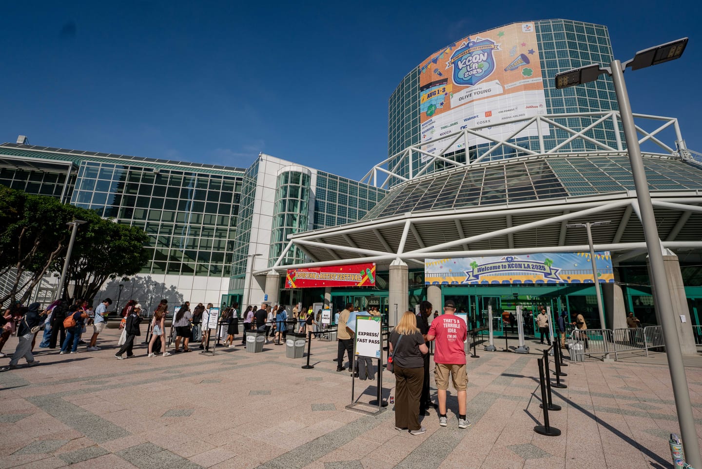 Fans queuing under the KCON LA 2025 banner on the globe facade of the Los Angeles Convention Center West Hall, beside an express fast pass lane