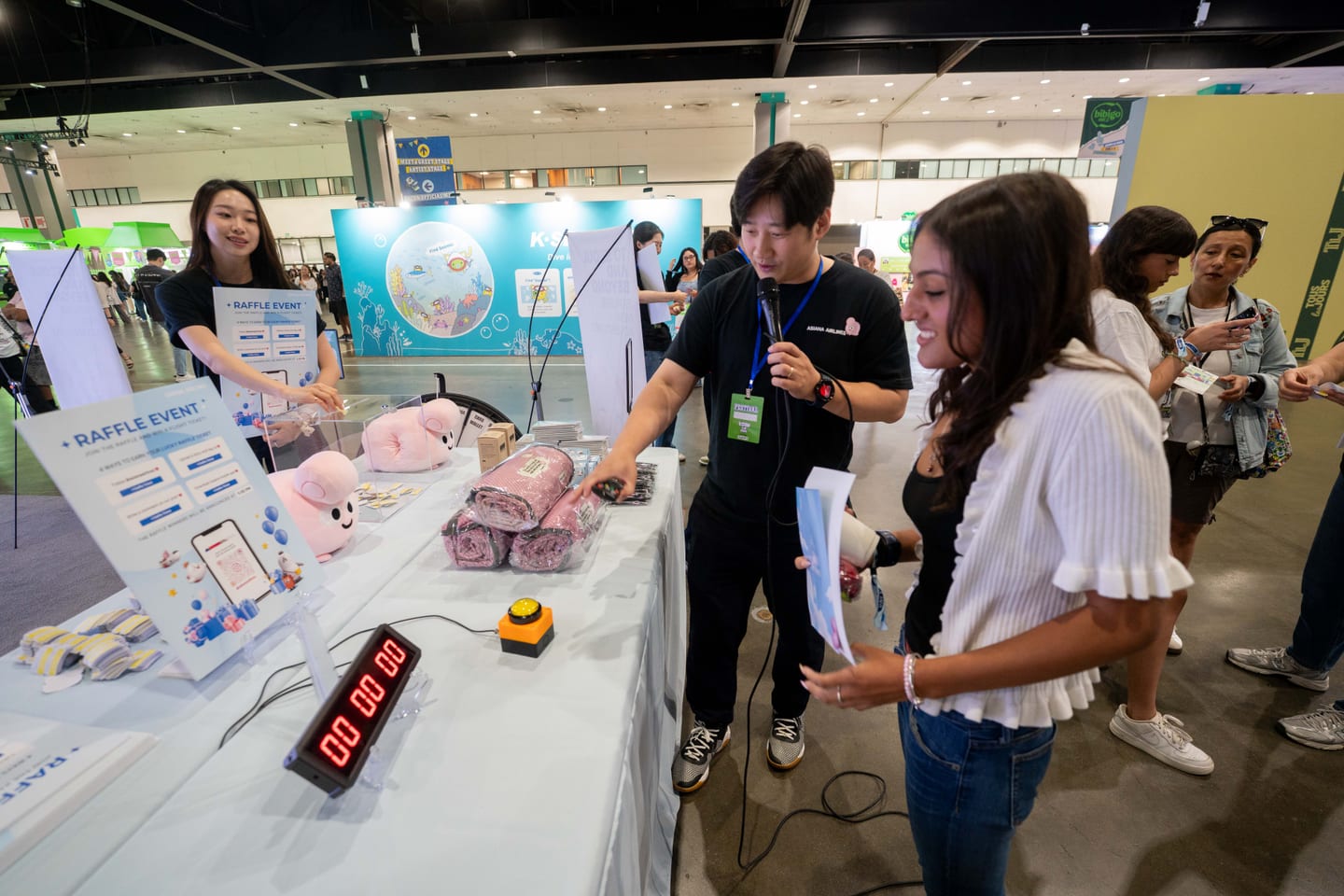 Staff running a timed raffle event at a KCON LA 2025 booth, with pink plush prizes and blankets stacked on the counter