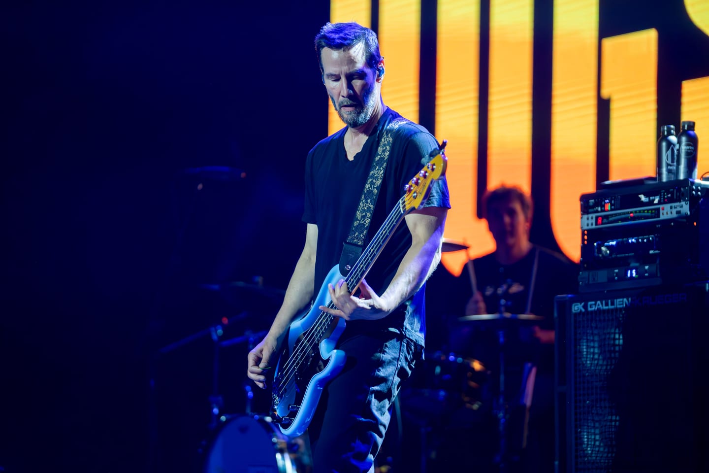 Keanu Reeves playing bass with his band Dogstar on a lit stage in front of a Los Angeles cityscape screen at IPW