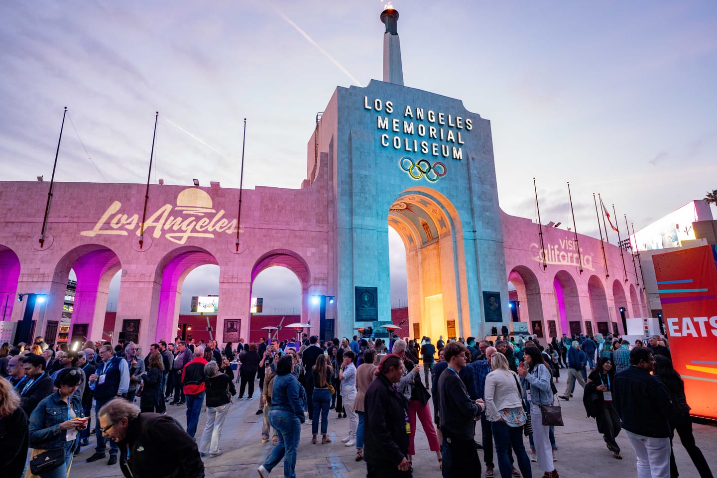 The exterior peristyle of the Los Angeles Memorial Coliseum lit pink at dusk with IPW delegates arriving