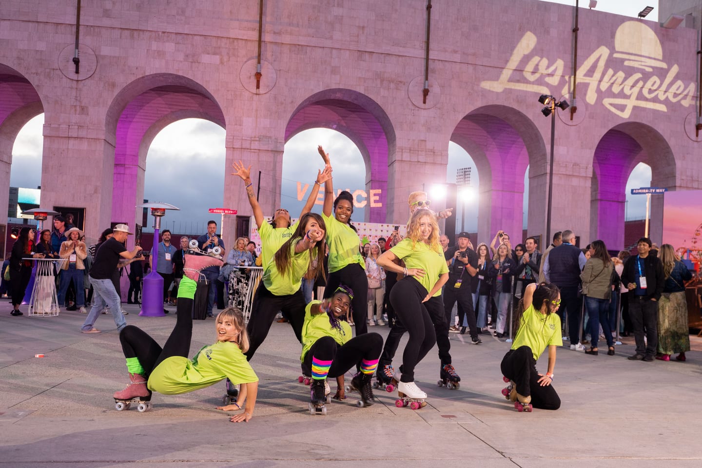 A roller-skating dance crew in bright green posing under the pink-lit arches of the Los Angeles Memorial Coliseum beneath a Los Angeles sign