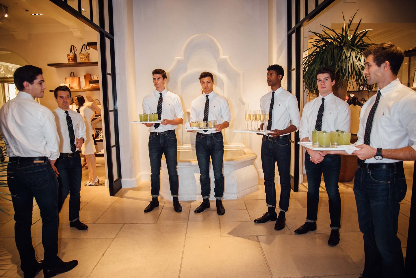 A row of servers in white shirts and ties holding welcome drink trays at an upscale retail boutique event
