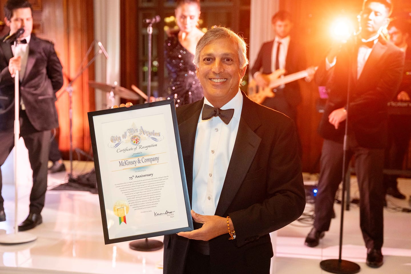 Executive holding a City of Los Angeles certificate at the McKinsey & Company 75th-anniversary gala, band performing behind