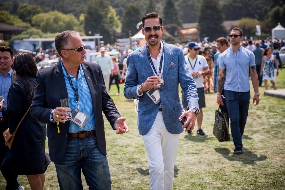 Well-dressed guests walking with drinks across a sunny festival lawn among tents at a Monterey Car Week event