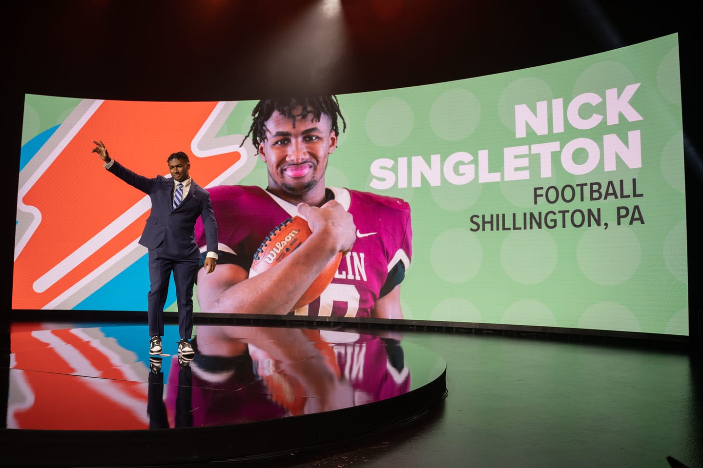 Football honoree Nick Singleton waving on stage in front of a curved LED wall showing his portrait at the Gatorade Athlete of the Year awards in Los Angeles