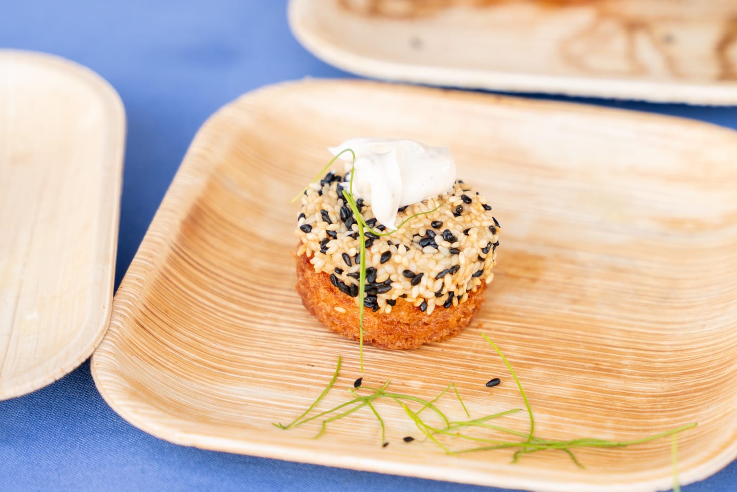 Close-up of a sesame-crusted crab cake topped with aioli and a microgreen on a palm-leaf plate at the Off the Hook seafood festival