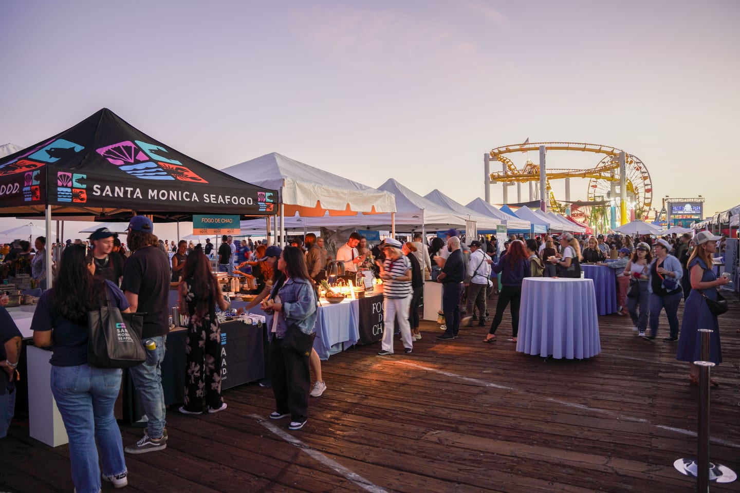 Vendor tents including Santa Monica Seafood and crowds at the Off the Hook seafood festival on the Santa Monica Pier at golden hour, the Pacific Park ferris wheel and roller coaster behind
