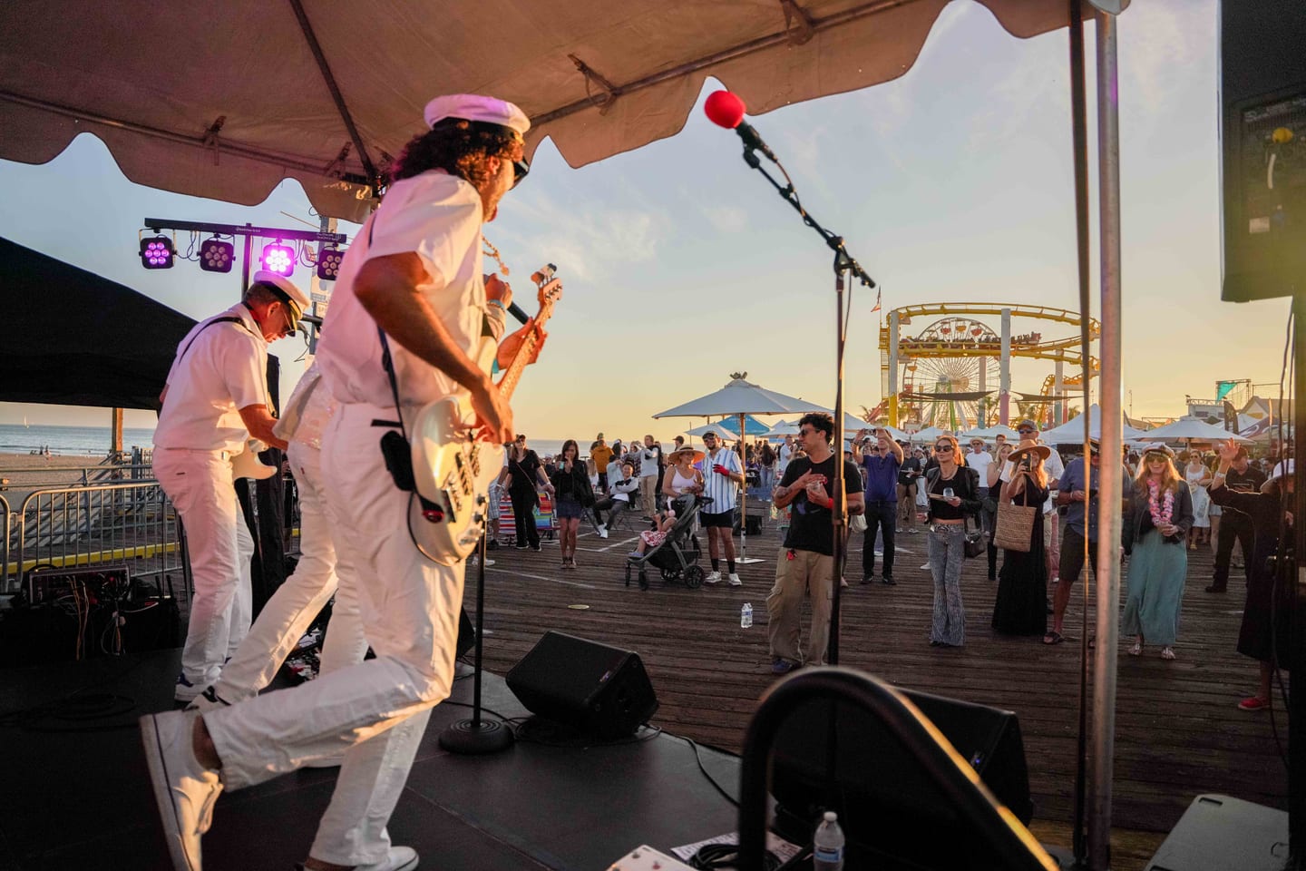 A band in white nautical outfits performing on an outdoor stage on the Santa Monica Pier at sunset, the Pacific Park ferris wheel and roller coaster behind, at the Off the Hook seafood festival