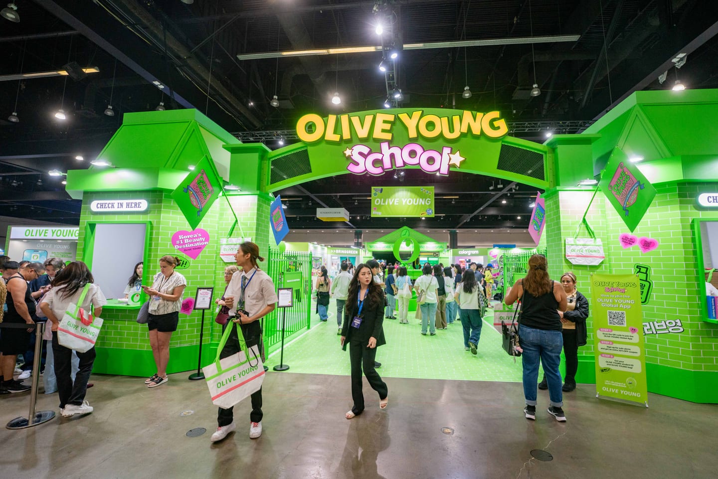 The bright green Olive Young School archway and check-in gates at KCON LA 2025, with staff handing out branded tote bags
