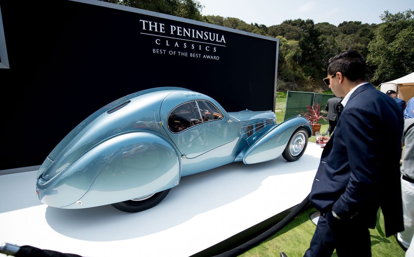 A rare pale-blue Bugatti Type 57SC Atlantic on a display platform at The Peninsula Classics Best of the Best Award, a guest admiring it on a concours lawn