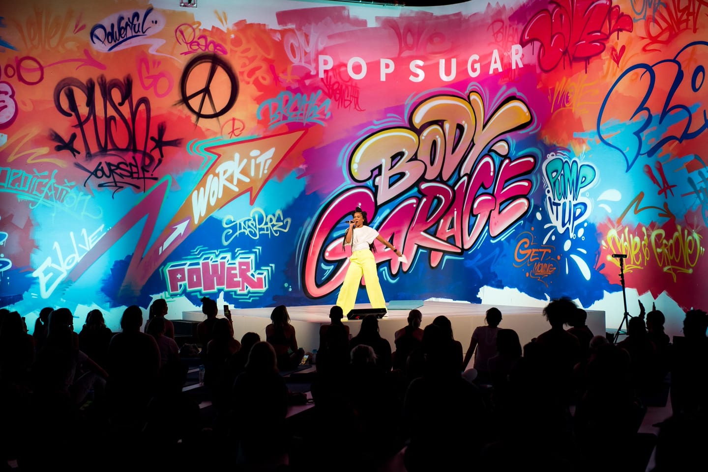 A host in yellow pants on stage in front of the giant graffiti PopSugar Body Garage mural, with a seated crowd silhouetted below
