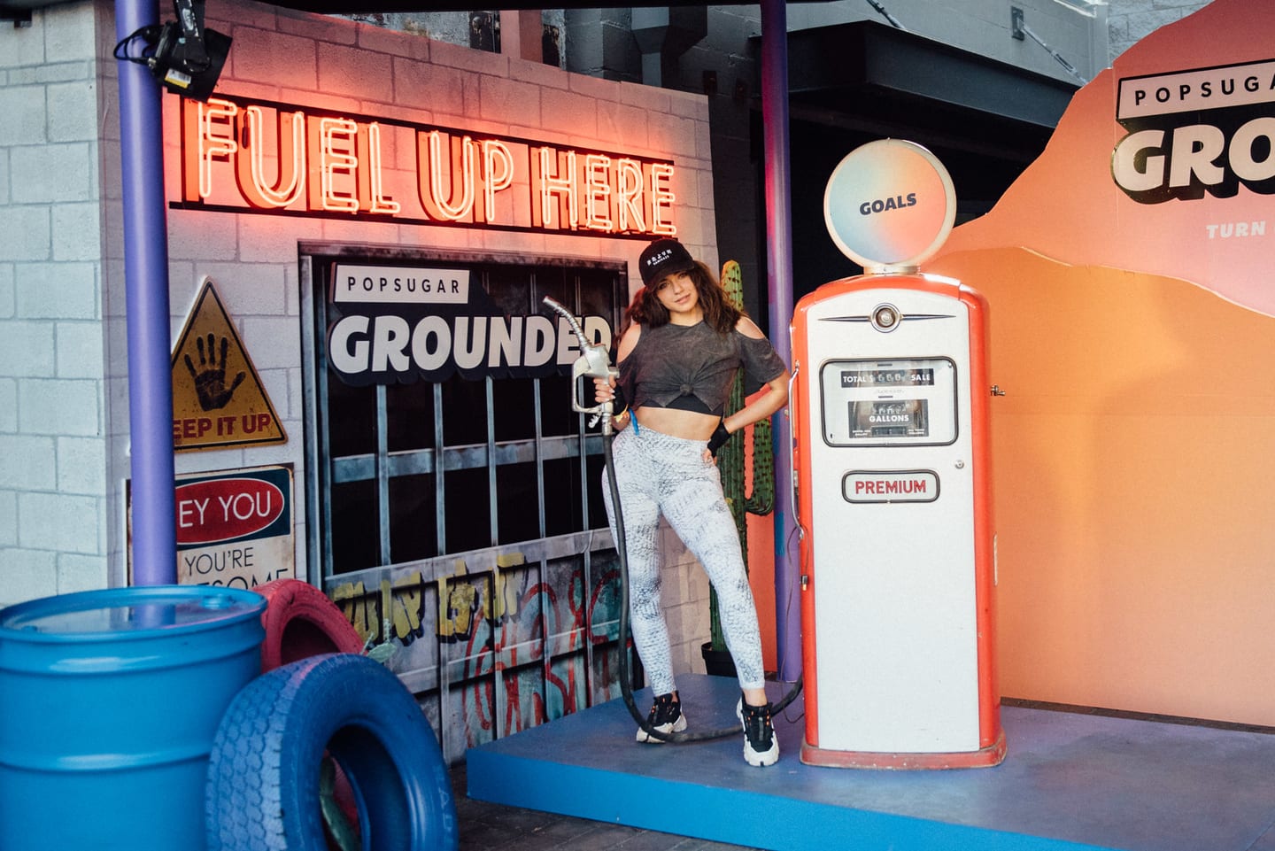 A guest posing with a vintage gas pump under a “Fuel Up Here” neon sign at the retro gas-station-themed PopSugar Grounded brand activation