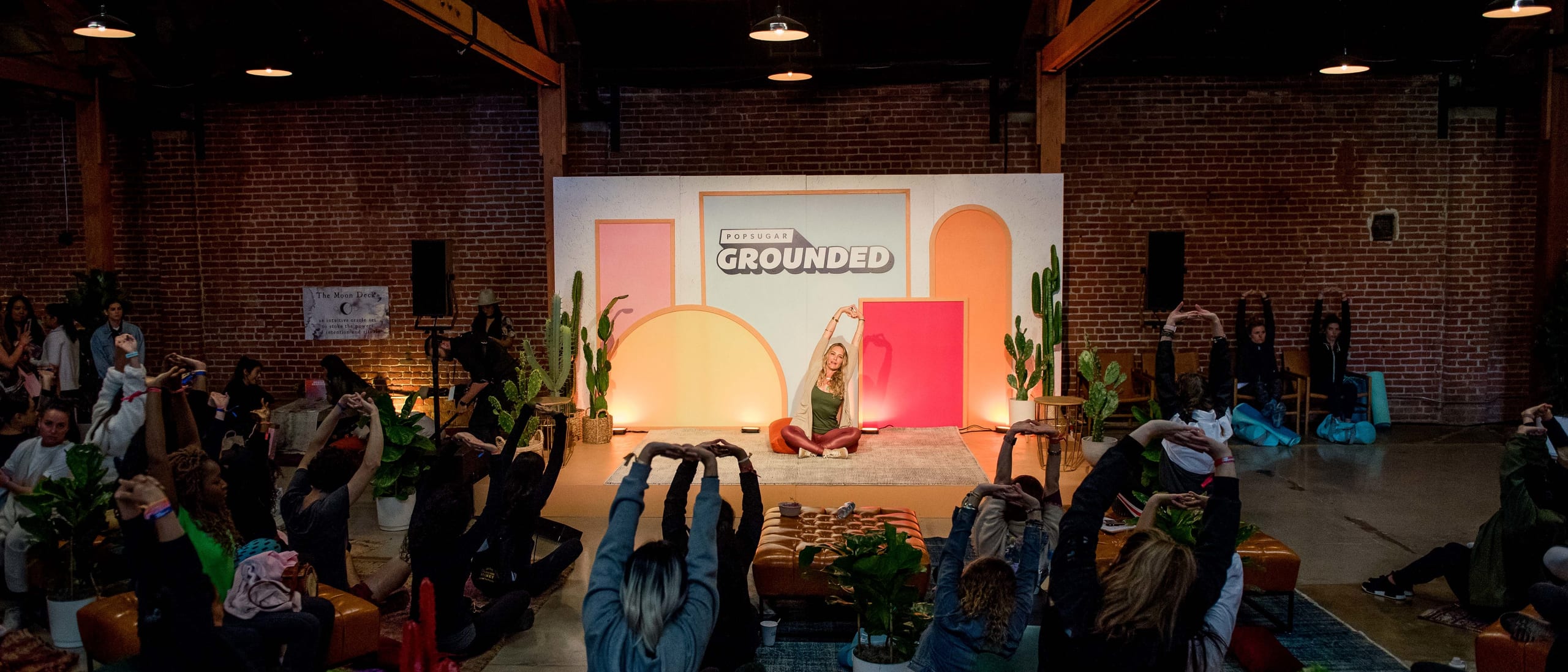 Wide view of guests following a guided stretch in front of the PopSugar Grounded stage inside a brick-walled Los Angeles venue