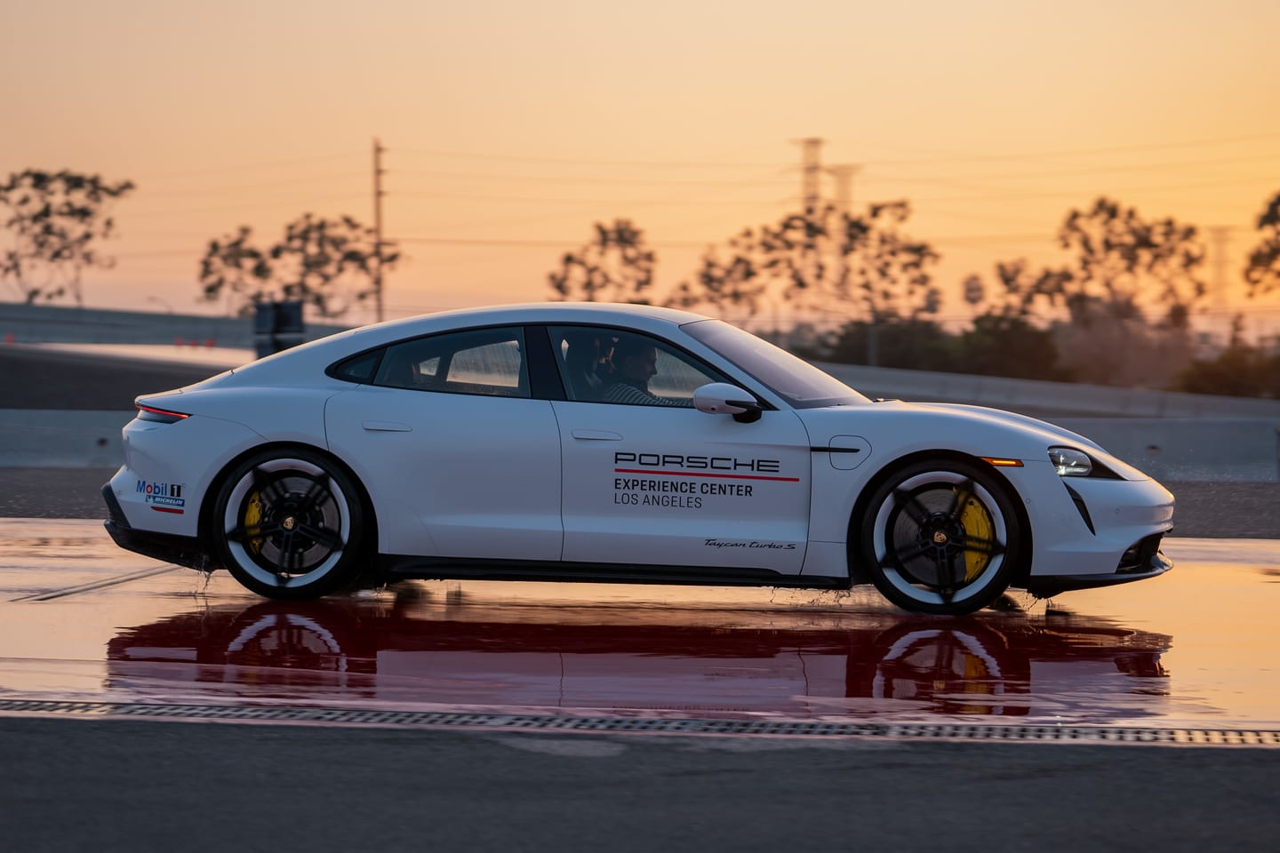 A white Porsche Taycan Turbo S driving on a wet handling track at sunset at the Porsche Experience Center Los Angeles, reflected on the water